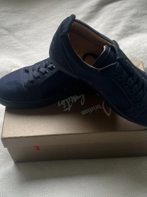 Marinblå sneakers från Christian Louboutin - Snygga marinblå sneakers från Christian Louboutin med klassisk röd sula och diskret logga på sidan. Skorna har snörning och är tillverkade i ett tygmaterial med detaljer i skinn. Perfekta för dig som vill ha något exklusivt och stilrent.