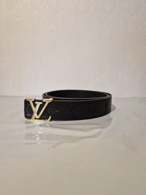 Louis Vuitton svart läderbälte - Snyggt svart bälte från Louis Vuitton i präglat läder med klassiskt LV-monogram längs hela bandet. Spännet är i guldmetall och har den ikoniska LV-loggan. Perfekt accessoar för att lyfta vilken outfit som helst.