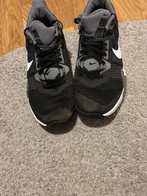 Svarta Nike sneakers med vit sula - Säljer ett par svarta Nike sneakers med vit sula och grå detaljer. Skorna har en stor vit Swoosh på sidan, mesh-överdel för bra andningsförmåga och snörning framtill. Perfekta för träning eller vardagsstil. Bekväm och sportig design.