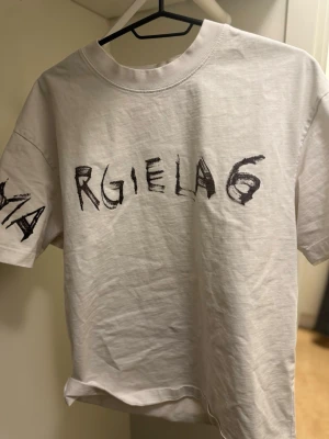 Vit Maison Margiela 6 t-shirt - Säljer en vit t-shirt från Maison Margiela med svart grafiskt tryck 'Margiela 6' på framsidan och ärmen. T-shirten har klassisk passform, rund halsringning och är gjord i mjuk bomull. Perfekt för dig som gillar streetwear och unika designer.