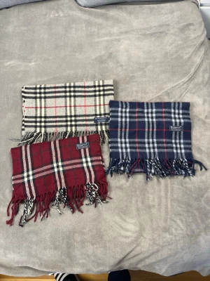 Rutiga halsdukar från Burberrys - Tre klassiska halsdukar från Burberrys i 100% lammull. En beige med svart och rött rutmönster, en vinröd med vitt och svart rutmönster samt en mörkblå med vitt och rött rutmönster. Alla har fransar i ändarna och den ikoniska Burberrys-loggan.