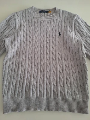  kabelstickad tröja Polo Ralph Lauren - Kabelstickad grå tröja från Polo Ralph Lauren med rund halsringning och klassisk broderad logga på bröstet. Tröjan har ribbade muddar vid ärmslut och nederkant. Perfekt för dig som gillar stilren och tidlös design.