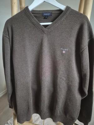 Brun stickad tröja från Gant, storlek L - Mörkbrun stickad tröja från Gant med V-ringning och diskret logga broderad på bröstet. Tröjan har långa ärmar, ribbade muddar. Tvättas i 40 grader.