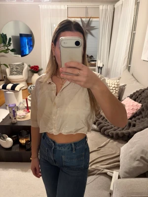 Beige croppad blus med knappar - Säljer en beige croppad blus med krage och knappar framtill. Blusen har en avslappnad passform och är tillverkad i ett tunt, lätt material som ger en snygg och luftig känsla. Perfekt att styla med höga jeans eller kjol.