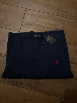 Mörkblå kabelstickad tröja Polo Ralph Lauren - Mörkblå kabelstickad tröja från Polo Ralph Lauren med rund halsringning och den klassiska röda logotypen broderad på bröstet. Tröjan har långa ärmar och ribbade muddar vid ärmslut och nederkant. Perfekt för en stilren och tidlös look.
