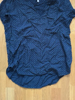 Mörkblå blus med småmönster Esprit - Snygg mörkblå blus från Esprit med små vita och vinröda prickmönster. Blusen har korta ärmar och rundad nederkant, tillverkad i ett lätt och luftigt tyg som känns skönt mot huden. Perfekt till jeans eller kjol för en avslappnad look.