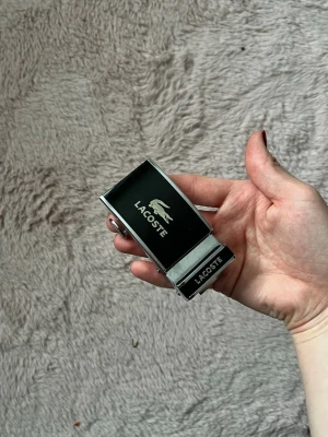 Lacoste metallspänne till bälte - Säljer ett stilrent bältesspänne från Lacoste i metall med svart front och den klassiska krokodilloggan. Spännet har en modern, fyrkantig form och passar perfekt till ett tyg- eller läderbälte för en clean look.