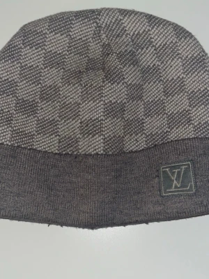 Grå Louis Vuitton mössa  - Snygg grå mössa från Louis Vuitton med klassiskt rutmönster och diskret LV-logga framtill. Tillverkad i mjuk ullmix som håller dig varm och stilren. Perfekt accessoar för dig som vill ha en exklusiv touch på din outfit.