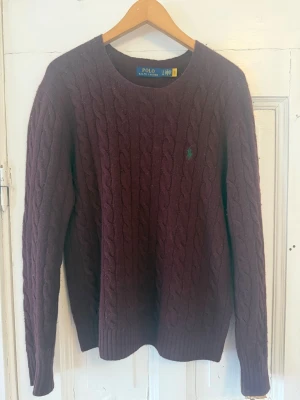 Lila kabelstickad tröja Polo Ralph Lauren - Kabelstickad tröja i mörklila från Polo Ralph lauren, med en kashmir och ull blend. En riktigt klassiker som passar rikitgt bra nu på vintern