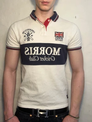 Vit Morris pikétröja med tryck - Vit pikétröja från Morris med mörkblått bröstband och tryck. Tröjan har korta ärmar, krage med röd insida och broderade detaljer som brittisk flagga och text. Klassisk passform och sportig look, perfekt till jeans.
