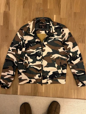 Camouflage vindjacka från Woolrich - Snygg vindjacka från Woolrich i klassiskt camouflage-mönster med dragkedja framtill och två fickor. Jackan har en krage och elastiska muddar vid ärmslut och midja. Perfekt för dig som vill ha en streetig look.