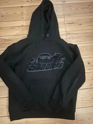 Svart Trapstar London Shooters hoodie - Säljer en svart hoodie från Trapstar med stor broderad 'London Shooters'-logga och pantermotiv på bröstet. Klassisk passform med huva och känguruficka framtill. Perfekt för dig som gillar streetwear och vill sticka ut med en unik design.