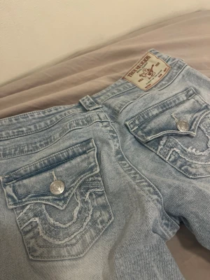 Ljusblå True Religion bootcut jeans - Säljer ett par ljusblå jeans från True Religion med ikoniska bakfickor och silverfärgade knappar. Jeansen har bootcut passform och klassisk femficksdesign. Materialet är denim i bomull och de har snygga slitningar på fickorna. Aldrig använt🥰