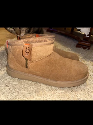 UGG Classic Ultra Mini Boot - Mysiga UGGs i färgen Chestnut i mjuk mocka med fluffigt foder. Klassisk rund tå och platt sula, perfekt för kyliga dagar. Dragkedja på sidan och UGG-logga på skaftet. Snygg och enkel design som passar till det mesta.