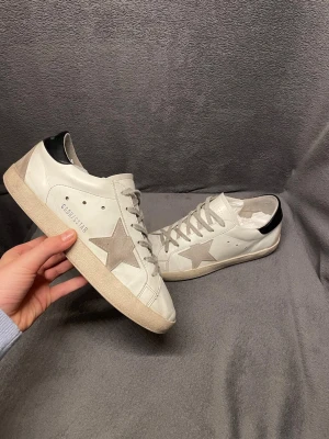Golden Goose  - Golden Goose sneakers i vitt skinn med grå mockadetaljer och ikonisk stjärna på sidan, i storlek 41. Skorna har används några gånger och har tecken på användning. Nypris 5800. OG är box och boxen medföljer vid köp. Perfekt för varma sommar dagar men också tidig höst och sen vår. Perfekt för dig som vill ha en unik stilren look till vardagen. Kan också gå med på byten.                                                               Skriv vid frågor, priset är inte hugget i sten🤝