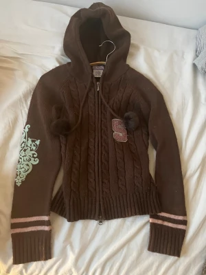 Y2K ,Brun stickad hoodie från tripple five soul - Mysig brun stickad hoodie från Triple Five Soul med kabelstickat mönster, dragkedja och luva med tofsar. Rosa och ljusblå detaljer på ärmarna samt ett broderat S på bröstet. Ribbstickade muddar och sportig collegekänsla.