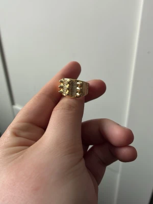 Guldfärgad chunky ring med nitar - Cool chunky ring i guldfärg med nitar och detaljerad struktur på ovansidan. Ringen är öppen baktill vilket gör den justerbar i storlek. Perfekt för dig som gillar statement-smycken och vill sticka ut lite extra.