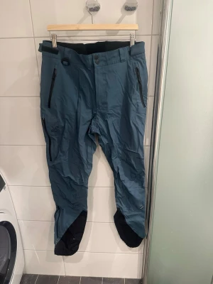 Cyclone 3L shell pants - Snygga blå täckbyxor revolutionrace med svarta förstärkningar vid bensluten och dragkedjor på fickorna. Perfekta för vinterns alla äventyr. Tillverkade i slitstarkt syntetmaterial och har en modern passform. Dessvärre finns en lagning i grenen som syns på bilderna, inget som syns när man bär dem. För män men jag har burit dem som tjej så passar båda