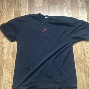 Svart Gymshark t-shirt med röd logga - Snygg svart t-shirt från Gymshark med liten röd logga på bröstet och stor GYMSHARK-text i rött på ryggen. Klassisk passform med rund hals och korta ärmar. Perfekt för dig som gillar sportig och enkel stil.