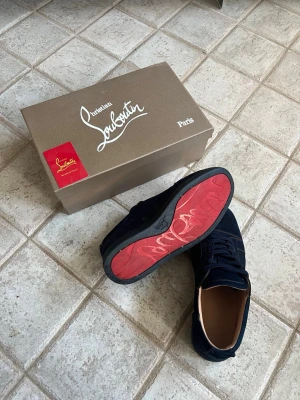 Christian Louboutin skor  - Snygga mörkblå sneakers från Christian Louboutin i mocka med klassisk röd sula och diskret broderad logga på hälen. Skorna har rund tå, snörning och en stilren design som sticker ut med den ikoniska röda undersidan. Har box. Någon fläck på moccan. Priset kan diskuteras 