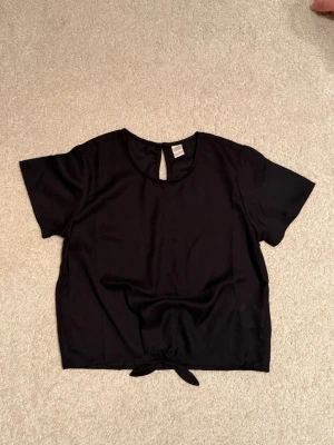 Svart festlig kortärmad blus med knyt. - En svart, enkel blus med korta ärmar och rund halsringning. Toppen har en liten öppning med knapp i nacken och knyt framtill vid midjan för en snygg detalj. Passar extra fint med ett svart linne under, då den är lite transparent i tyget.