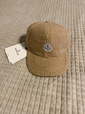 Beige/karamell teddykeps från Moncler - Snygg beige keps från Moncler i mjukt teddy-material. Kepsen har klassisk form med böjd skärm och Monclers ikoniska logga framtill. Perfekt accessoar för att lyfta din outfit med en trendig och mysig touch.