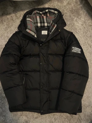 Svart dunjacka från Burberry - Snygg svart dunjacka från Burberry med huva och rutigt innerfoder. Jackan har tryckknappar, dragkedja och logga på ärmen. Perfekt för kalla dagar och har en klassisk pufferstil med quiltade sektioner.