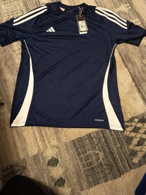 Adidas Tiro24 blå fotbollströja XL barn - Mörkblå sporttröja från Adidas med vita detaljer på axlar och sidor. Tillverkad i lätt och ventilerande material med Aeroready-teknologi. Perfekt för fotbollsträning eller annan sport. Klassisk Adidas-logga på bröstet och snygg passform.