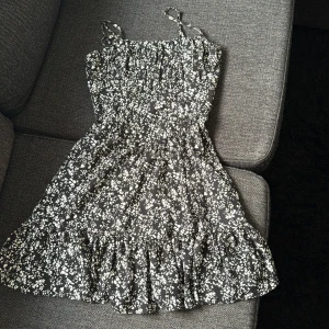 Svart blommig kortklänning NA-KDxPAMELA - Svart klänning med vita blommor från Pamela x NA-KD. Modellen är kort med volangkant nertill och smala och justerbara axelband. Klänningen har dubbel tyg. (Nedkommer fläckar pgd av brun utan sol, enbart på insidan)