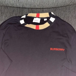 Svart Burberry sweatshirt med röd logga - Burberry sweatshirt Svart långärmad sweatshirt med röd broderad Burberry-logga på bröstet. Klassiska Burberry-ränder i kragen (beige, svart, vitt & rött). Köpt i Burberry-butik i USA. Säljes då den blivit för liten. 