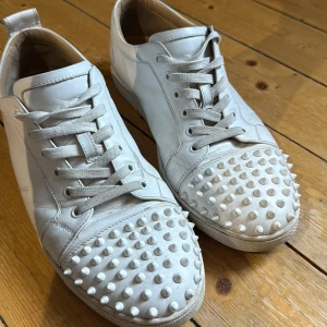 Vita louboutins - Säljer ett par vita sneakers från Christian Louboutin i skinn med coola nitar på tån och den klassiska röda sulan. Skorna har snörning och en rund tå. Perfekta för dig som vill sticka ut med din stil. Skick 6-7/10 såklart är dom även äkta som man kan se under sulan 