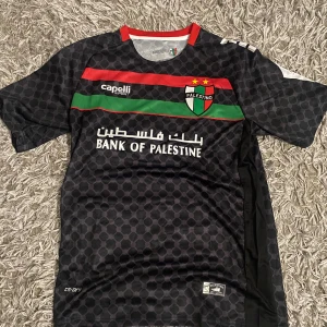 Svart Palestino matchtröja Capelli - Säljer en svart Palestino fotbollströja från Capelli Sport med korta ärmar. Tröjan har ett hexagonmönster, röd och grön rand över bröstet, klubbmärke med två gula stjärnor och tryck från Bank of Palestine. Materialet är lätt polyester, perfekt för träning eller match.