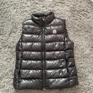 Svart dunväst från Moncler - Snygg svart dunväst från Moncler med glansig finish och klassisk quiltad design. Västen har hög krage, dragkedja framtill och Moncler-logga på bröstet. Den är storlek xs den har en litet hål som man ser på bilden och den har ingen tag där inne den har åkt bort men den är äkta är 99procent på det 