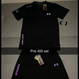 Svart Under Armour Set  - Svart t-shirt från Under Armour med färgglada loggor och tryck på bröstet och ärmen. T-shirten har korta ärmar och rund hals, perfekt för gymmet eller sport. Materialet är lätt och andas, vilket gör den skön att ha på sig under träning.