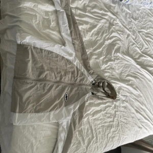 Beige och vit vindjacka Corteiz S - Säljer en beige och vit vindjacka från Corteiz Guerillaz med huva och dragkedja. Jackan har en clean tvåfärgad design, svartvit logga på bröstet och meshfoder på insidan. Perfekt för dig som gillar streetwear och vill sticka ut med en unik jacka.