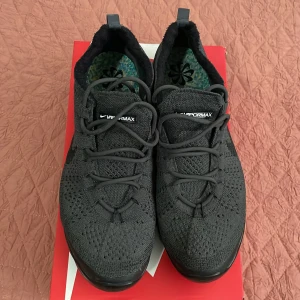 Nike Vapormax svarta sneakers - Nya, knappt använda svarta Nike Vapormax sneakers med stickad ovandel i flyknit-material och synlig luftsula. Skorna har rund tå, snörning och diskret Nike-logga på sidan. Perfekta för dig som gillar en sportig och modern look med maximal komfort.