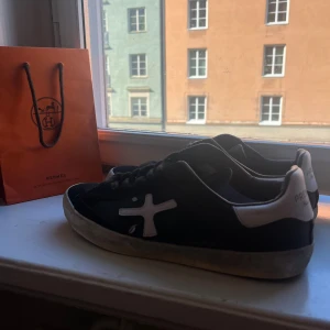 Premiata sneakers svart och vit - Svarta sneakers från Premiata med vit logga på sidan och vit häl med texten 'Premiata'. Skorna har snörning och platt sula, samt en rund tå. Materialet ser ut att vara skinn och syntet. Snygg kontrast mellan svart och vitt, perfekt för dig som gillar stilrena sneakers.