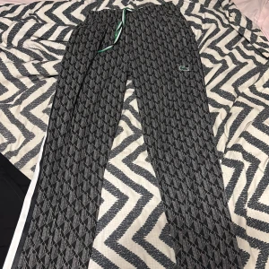 lacoste mjukis - Snygga svarta mjukisbyxor från Lacoste med geometriskt mönster och vit sidoremsa. Byxorna har grön snörning i midjan och den klassiska Lacoste-loggan broderad på benet. Perfekta för chill eller streetwear-stil.