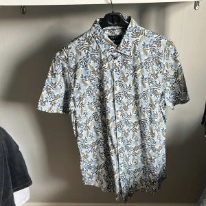 Blommig kortärmad skjorta från IVEO - Snygg kortärmad skjorta från IVEO i slim fit-modell. Skjortan har ett blommigt mönster i blått, brunt och vitt med klassisk krage och knappar framtill. Perfekt för dig som vill sticka ut med en fräsch och somrig look.