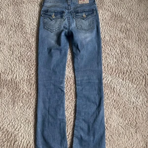 Blå bootcut jeans från True Religion - Snygga blå jeans från True Religion med bootcut passform, storlek 26. Jeansen har klassiska kontrastsömmar, dekorativa ficklock med knappar bak och ikonisk logga på bakfickan. Tillverkade i mjukt denim med lätt slitning.💗