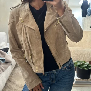 Beige mocka bikerjacka med guldiga detaljer - Snygg beige bikerjacka i mocka med guldiga dragkedjor och knappar. Långärmad och figurnära passform. Det är storlek L på den men jag har xs/s i vanliga fall så den är väldigt liten i modellen 