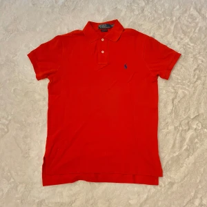 Röd pikétröja från Polo Ralph Lauren S - Riktigt snygg röd pikétröja från Polo Ralph Lauren med klassisk krage, två knappar och blå broderad logga på bröstet. Tröjan är i bomull och har en custom fit som sitter schysst på kroppen. Perfekt för dig som gillar stilren och tidlös design.