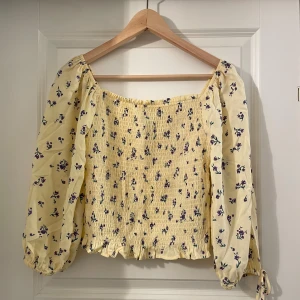 Gul blommig blus  - Supersöt gul blus med små lila blommor och smockad kropp. Vida långa ärmar med resår och knyt vid ärmslut. Fyrkantig ringning och volangkant nertill ger en romantisk vibe. Perfekt för vår och sommar!