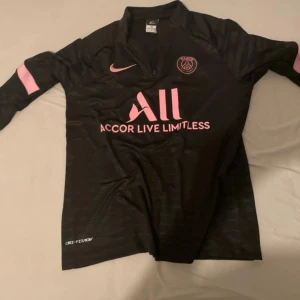 Svart PSG långärmad fotbollströja Nike S - Svart långärmad PSG fotbollströja från Nike med rosa detaljer och klubbmärke på bröstet. Tröjan har Dri-FIT-material som andas och rosa Nike-logga samt sponsortryck på framsidan. Perfekt för dig som älskar Paris Saint-Germain och snygg sportstil.
