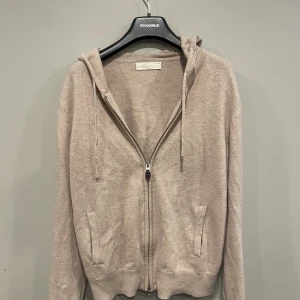 Beige hoodie med dragkedja från Sayless - Mysig beige hoodie från Sayless med dragkedja framtill, huva med snörning och två fickor. Tillverkad i mjukt stickat material, perfekt för lager-på-lager. Långärmad och med ribbade muddar för extra komfort.