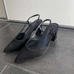 Svarta pumps med spetstå - Snygga svarta pumps från Anna Field med spetsig tå och bred klack. Skorna har en öppen häl med smala remmar och är tillverkade i mocka för en lyxig känsla. Skorna är i storlek 38