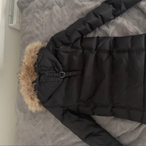 Svart pufferjacka med päls från Hollies - Säljer en svart pufferjacka i storlek xs från Hollies med snygg fuskpäls på huvan och flera praktiska fickor framtill. Jackan har långa ärmar, dragkedja och knappar, samt coola detaljer som spänne vid huvan och orange piping på insidan. Perfekt för kalla dagar!