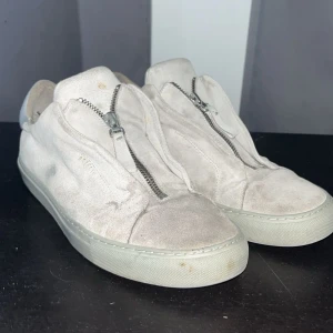 Ljusgrå sneakers från Axel Arigato - Snygga ljusgrå sneakers från Axel Arigato i mocka med dragkedja framtill istället för klassisk snörning. Skorna har rund tå, platt sula och stilren design. Perfekta för dig som gillar minimalistisk stil och vill ha något unikt på fötterna.