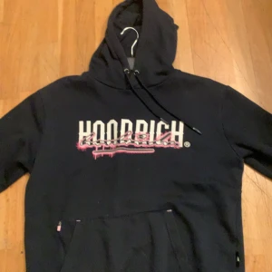 Mörkblå Hoodrich hoodie - Mörkblå hoodie från Hoodrich med stor vit och rosa logga framtill och text på ryggen. Hoodien har huva med snörning och en stor magficka. Snygga detaljer med rosa och vitt tryck som ger streetkänsla. Perfekt för dig som gillar urban stil.