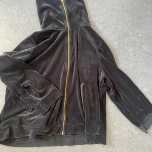 Svart velour hoodie med guldzip - Mjuk och skön svart hoodie i velour med guldig dragkedja framtill. Tröjan har hög krage, långa ärmar och två fickor på sidorna. Perfekt för chill dagar eller när du vill ha en extra cozy vibe.